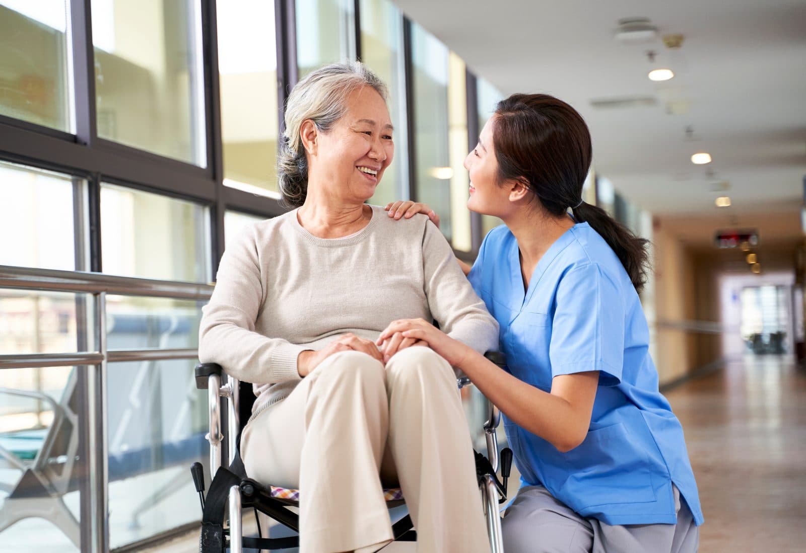 A smiling older woman in a wheelchair holding hands with a carer who kneels beside her
